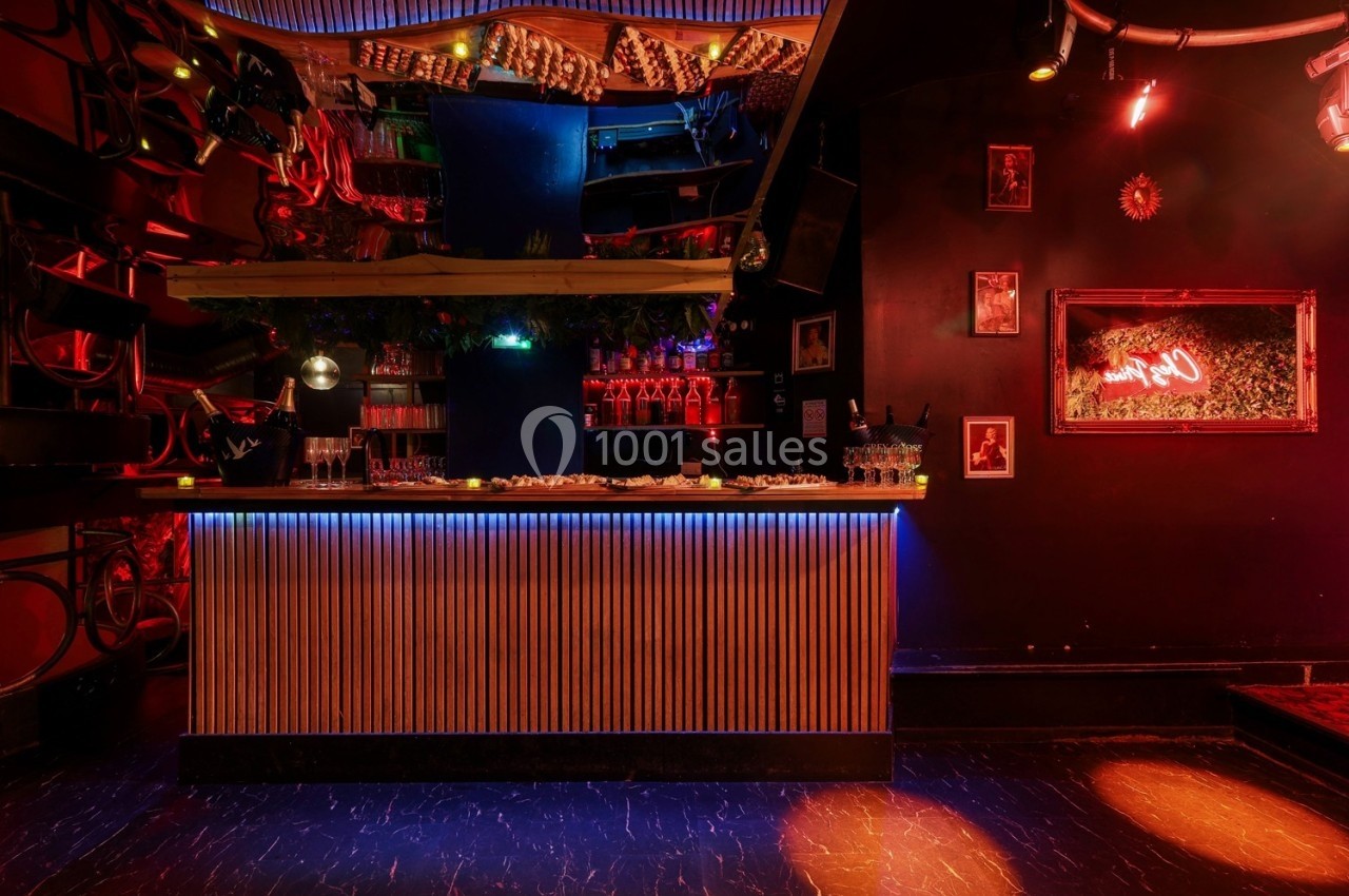 Bar avec comptoir éclairé, décorations lumineuses, murs sombres ornés de cadres et ambiance tamisée.