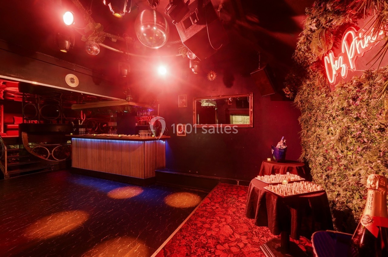 Salle de fête avec éclairage rouge, bar illuminé, miroir mural et décoration végétale sur un mur.