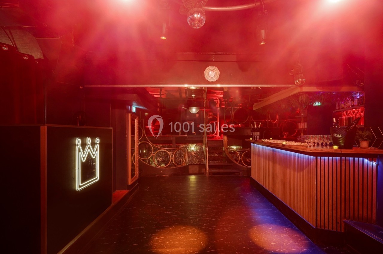Intérieur d'une discothèque avec éclairage rouge, bar éclairé et logo lumineux en forme de couronne sur un mur.