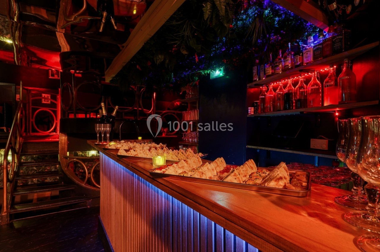 Plateau de toasts variés sur un comptoir en bois éclairé, entouré de bouteilles et de verres dans une ambiance tamisée.