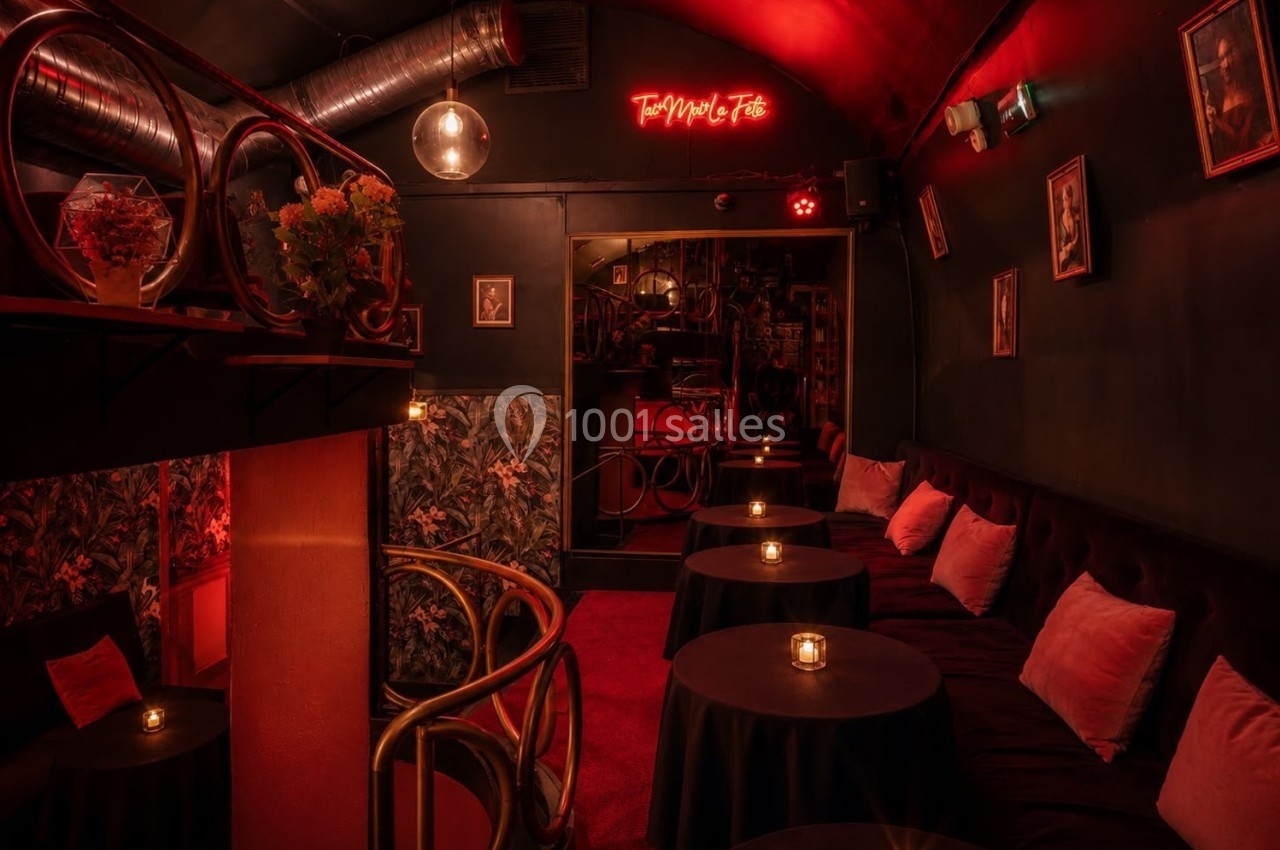 Salle intimiste éclairée par une lumière rouge tamisée, avec tables rondes, bougies et décorations murales.