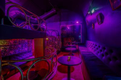 Salle sombre avec éclairage rouge, tables rondes, banquettes noires et décorations murales encadrées.