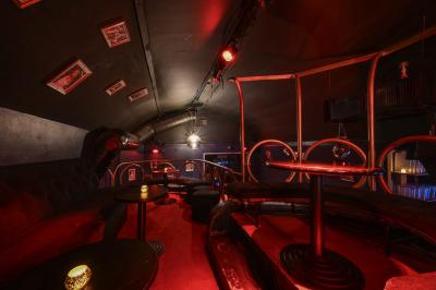 Salle sombre avec éclairage rouge, tables rondes, banquettes noires et décorations murales encadrées.