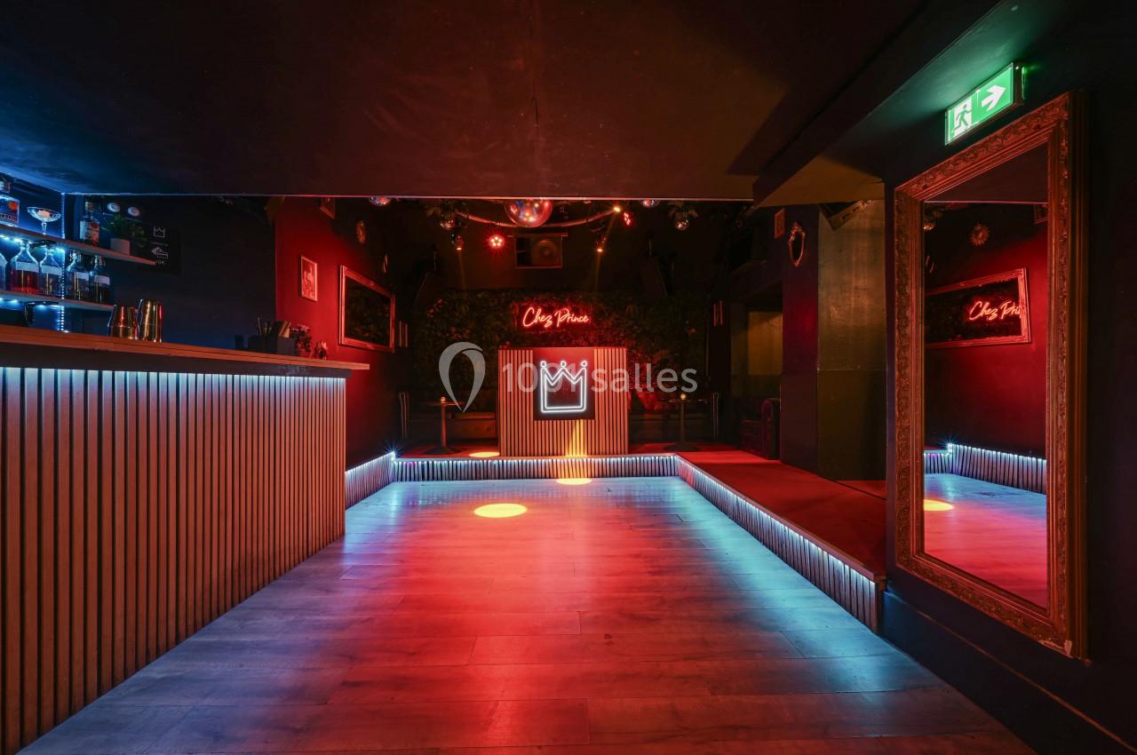 Salle de danse vide avec éclairage coloré, bar à gauche, miroir à droite et enseigne lumineuse en fond.