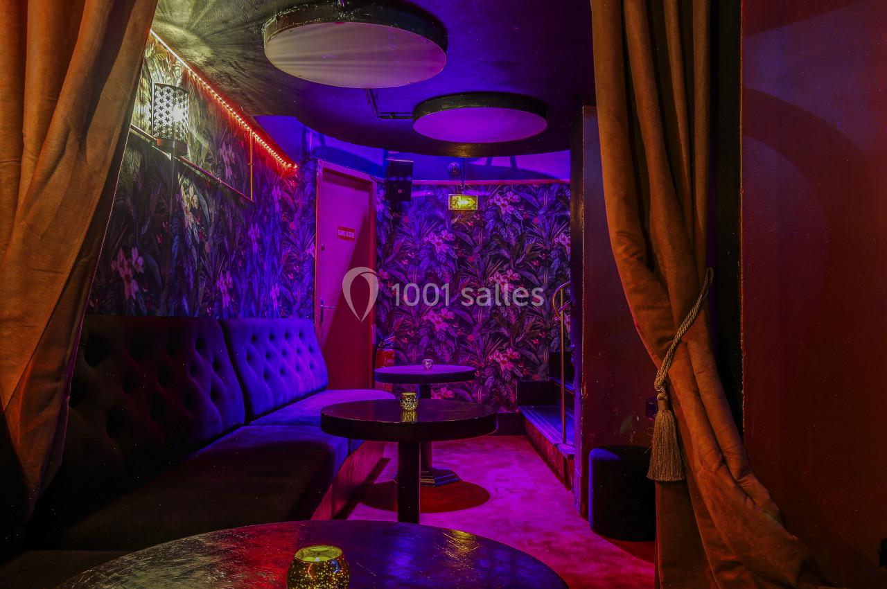 Salle intimiste avec banquettes noires, tables basses, rideaux et éclairage tamisé aux tons rouges et violets.