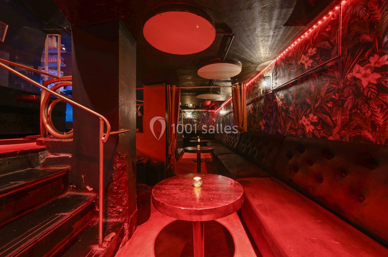 Salle de bar avec éclairage tamisé rouge, banquettes noires, tables rondes et murs décorés de motifs tropicaux.