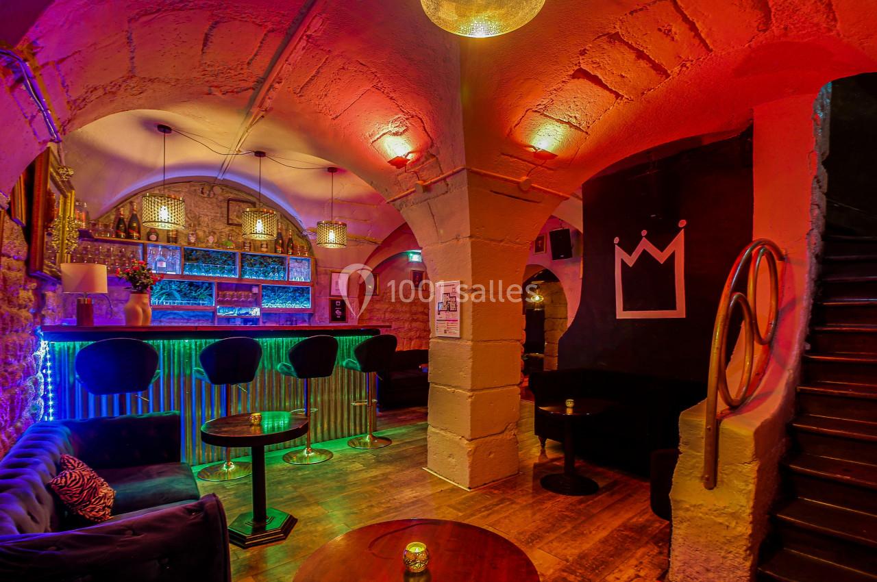 Intérieur d'un bar avec éclairage coloré, comptoir illuminé, tables basses, fauteuils et murs en pierre apparente.