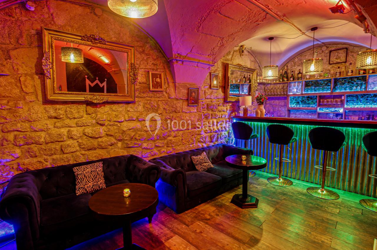 Intérieur d'un bar avec murs en pierre, canapés, tables basses, comptoir lumineux et éclairage coloré.