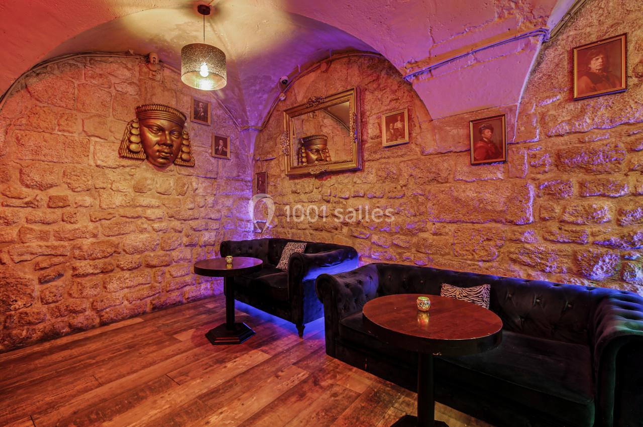 Salle voûtée en pierre avec canapés noirs, tables basses, éclairage tamisé et décorations murales.