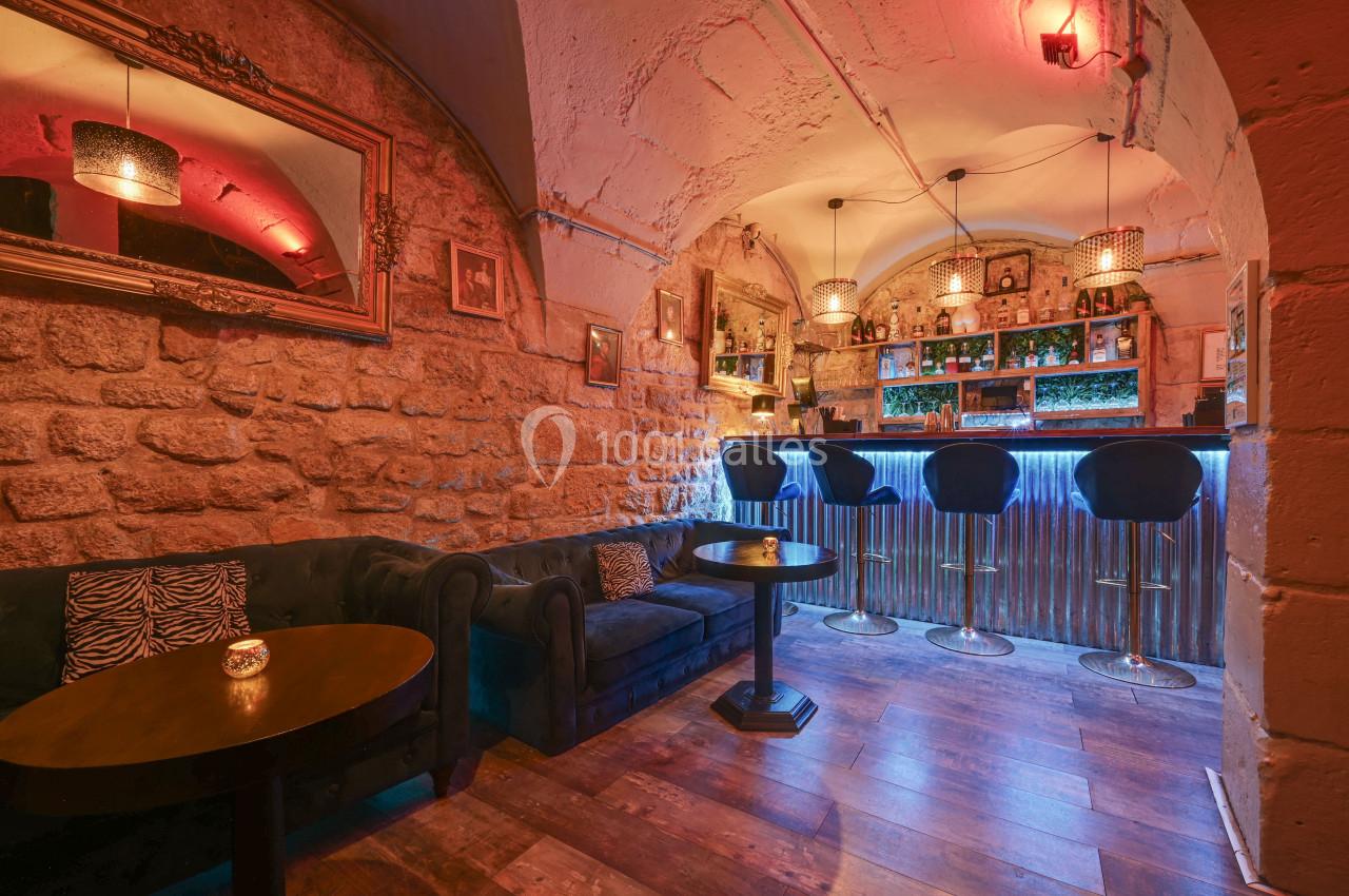 Intérieur d'un bar avec murs en pierre, canapé, tables basses, comptoir éclairé et tabourets hauts.