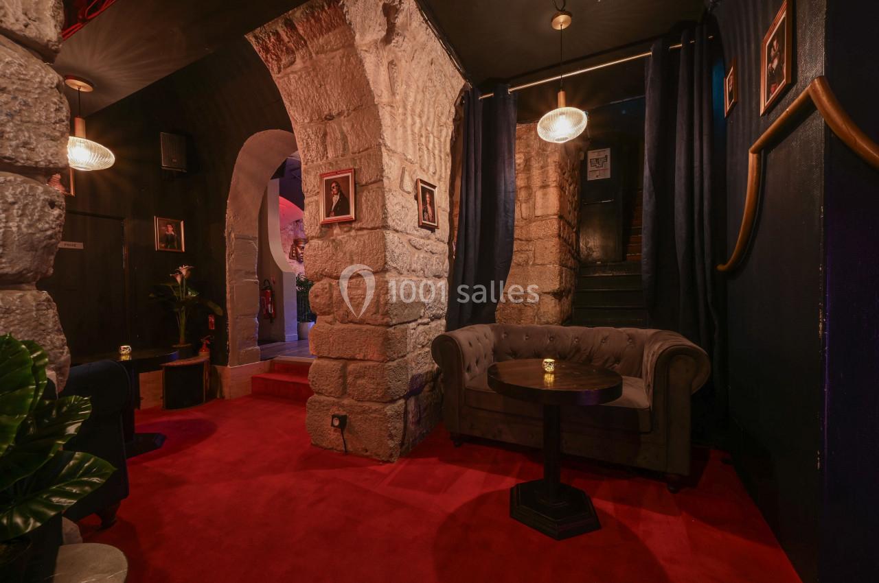 Espace intérieur avec murs en pierre, canapé gris, table basse, rideaux sombres et éclairage tamisé.