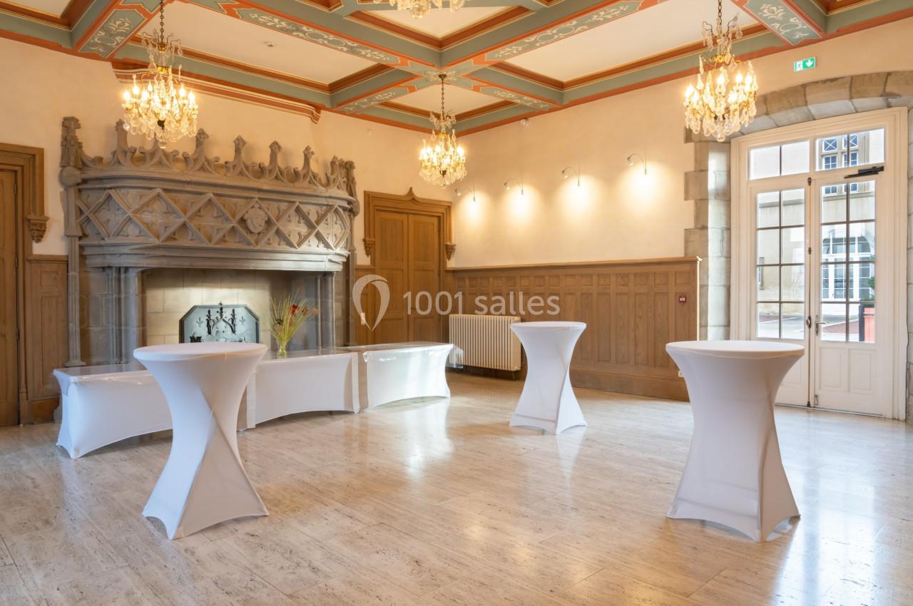 Salle élégante avec cheminée en pierre, tables hautes drapées de blanc et lustres suspendus au plafond décoré.