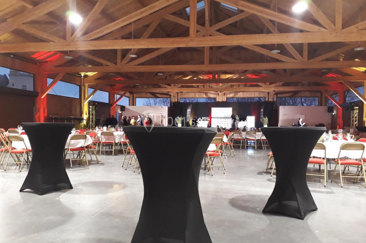 Salle spacieuse avec charpente en bois, tables hautes noires et chaises disposées pour un événement.