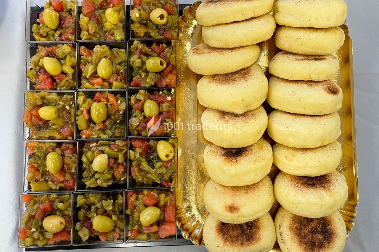 Plateau avec des mini bols de salade de légumes garnis d'olives vertes et des pains ronds dorés empilés.