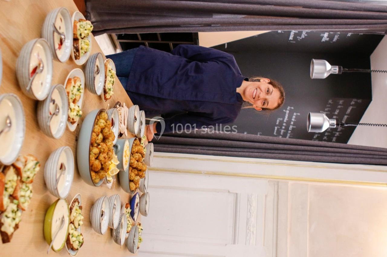 Une personne souriante en tenue de cuisine présente des plats variés disposés sur une table en bois.