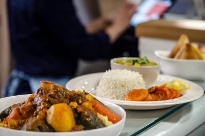 Assiette de couscous avec légumes et viande, accompagnée de riz, carottes et sauce, en arrière-plan une personne floue.