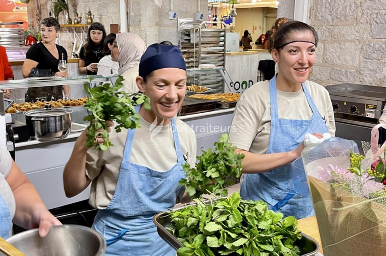 Deux cuisinières souriantes en tabliers bleus tiennent des herbes fraîches dans une cuisine animée.