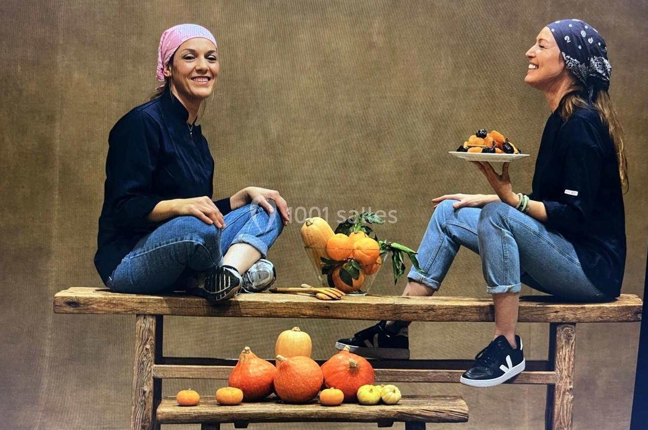 Deux femmes assises sur une table en bois, entourées de courges et de fruits, souriant dans un décor neutre.