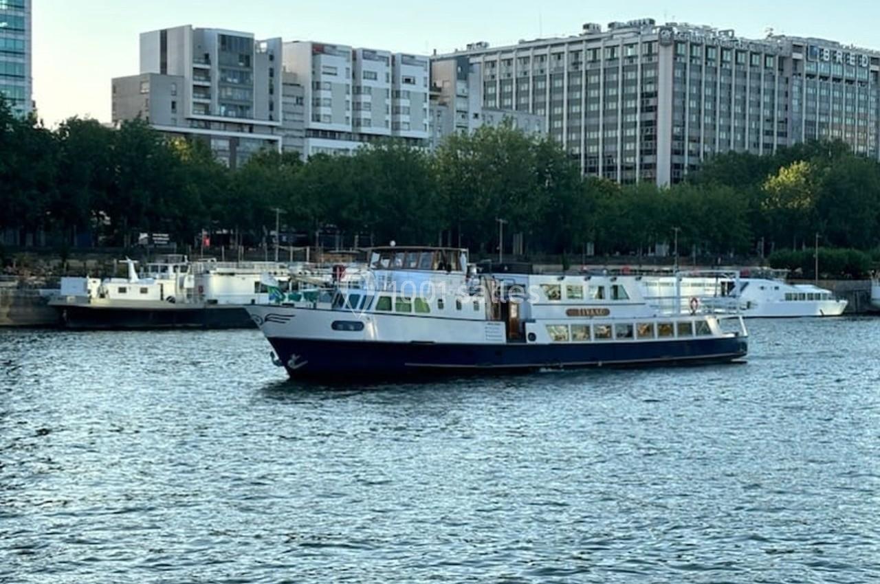 Bateau de croisière naviguant sur une rivière, avec des immeubles modernes et des arbres en arrière-plan.