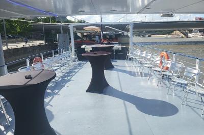 Pont supérieur d'un bateau aménagé avec des tables hautes et des chaises, amarré près d'une rive sous un ciel dégagé.