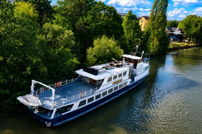 Un capitaine en uniforme utilise une radio dans la cabine d'un bateau, avec vue sur une rivière bordée d'arbres.