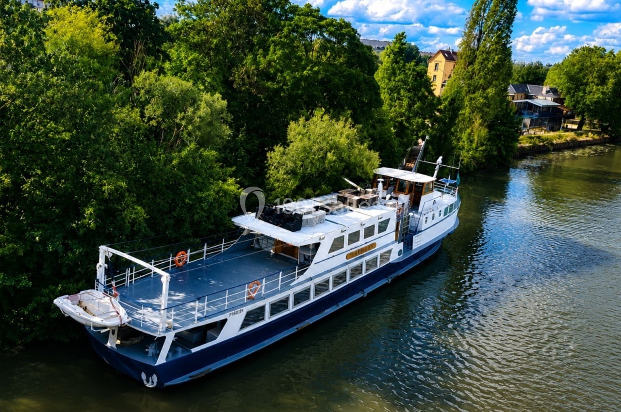 Bateau blanc et bleu amarré sur une rivière bordée d'arbres et de maisons sous un ciel partiellement nuageux.