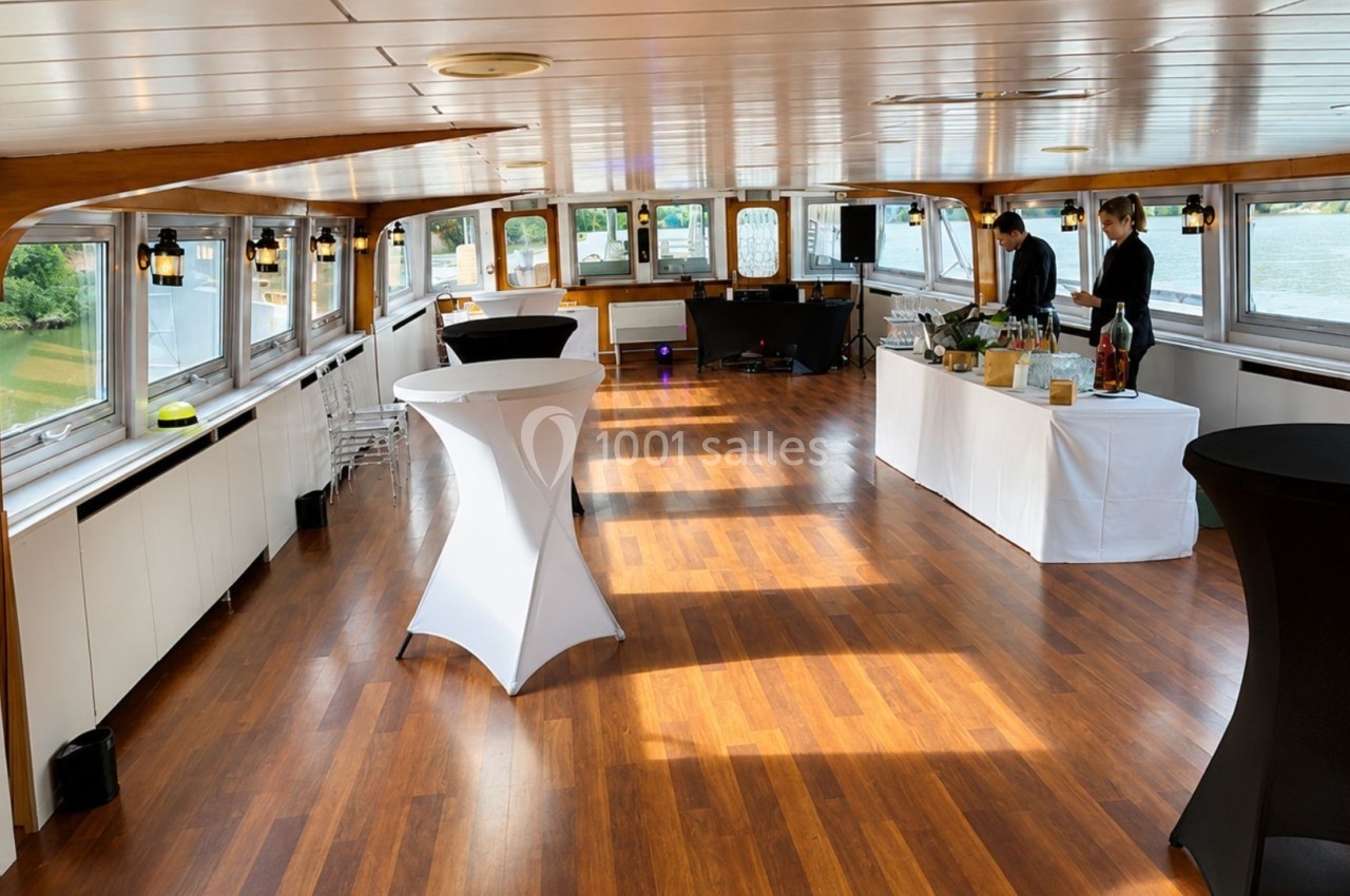 Salle lumineuse avec parquet en bois, tables hautes et buffet préparé, située sur un bateau avec vue sur l'extérieur.