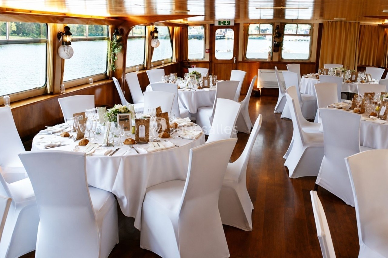 Salle de réception sur un bateau avec des tables rondes dressées, nappes blanches et vue sur l'eau par les fenêtres.