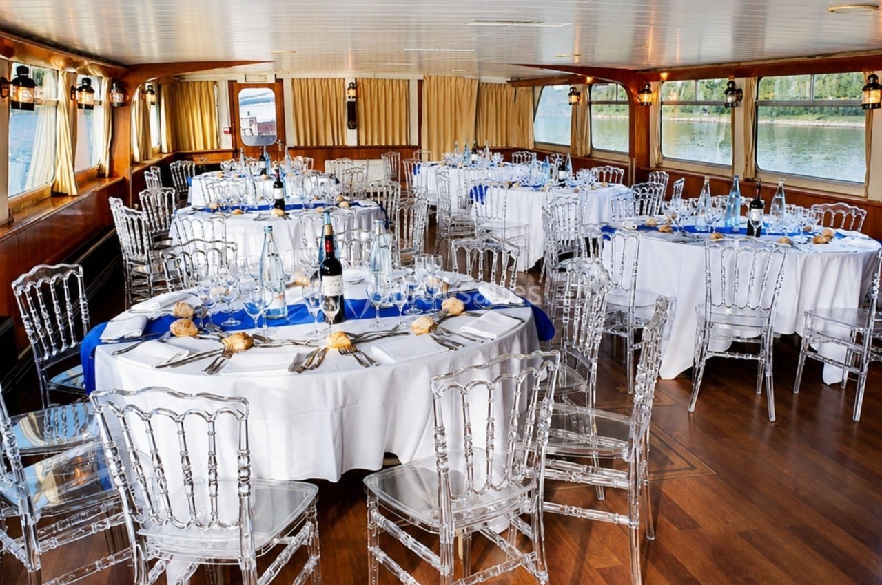 Salle de réception élégante sur un bateau, avec tables rondes dressées, chaises transparentes et vue sur l'eau.