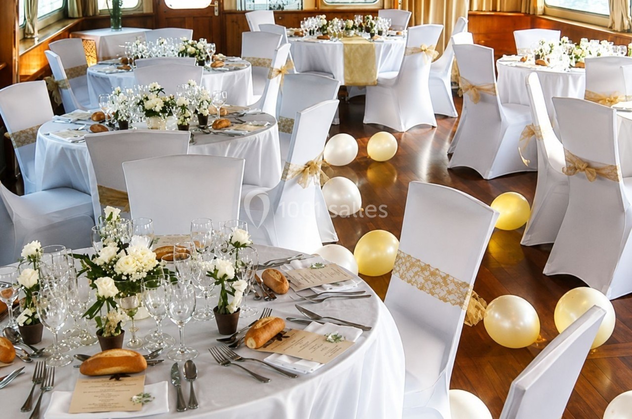 Salle décorée pour un événement avec des tables rondes dressées, nappes blanches, fleurs et ballons dorés au sol.