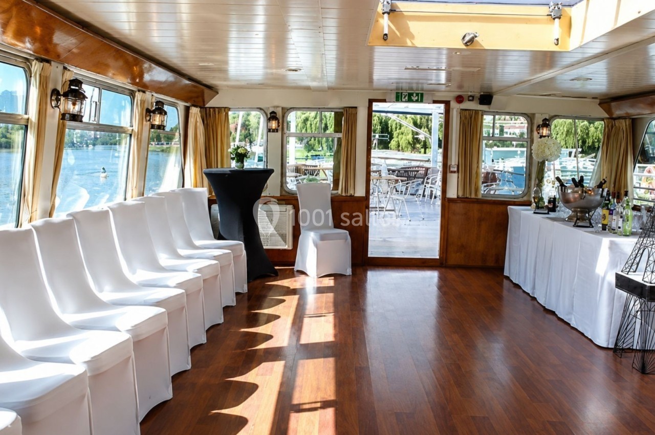 Intérieur d'un bateau avec des chaises blanches alignées, un sol en bois et une vue sur l'eau par les fenêtres.