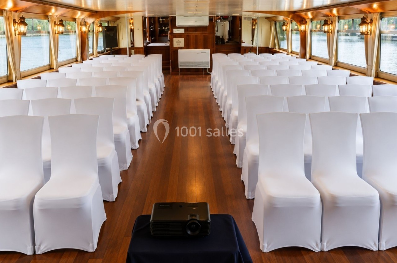 Salle de réception sur un bateau avec rangées de chaises blanches alignées et un projecteur au premier plan.