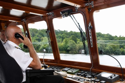 Un capitaine en uniforme utilise une radio dans la cabine d'un bateau, avec vue sur une rivière bordée d'arbres.