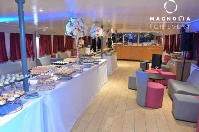 Salle de réception sur une péniche avec tables rondes dressées, rideaux rouges et buffet de desserts au premier plan.
