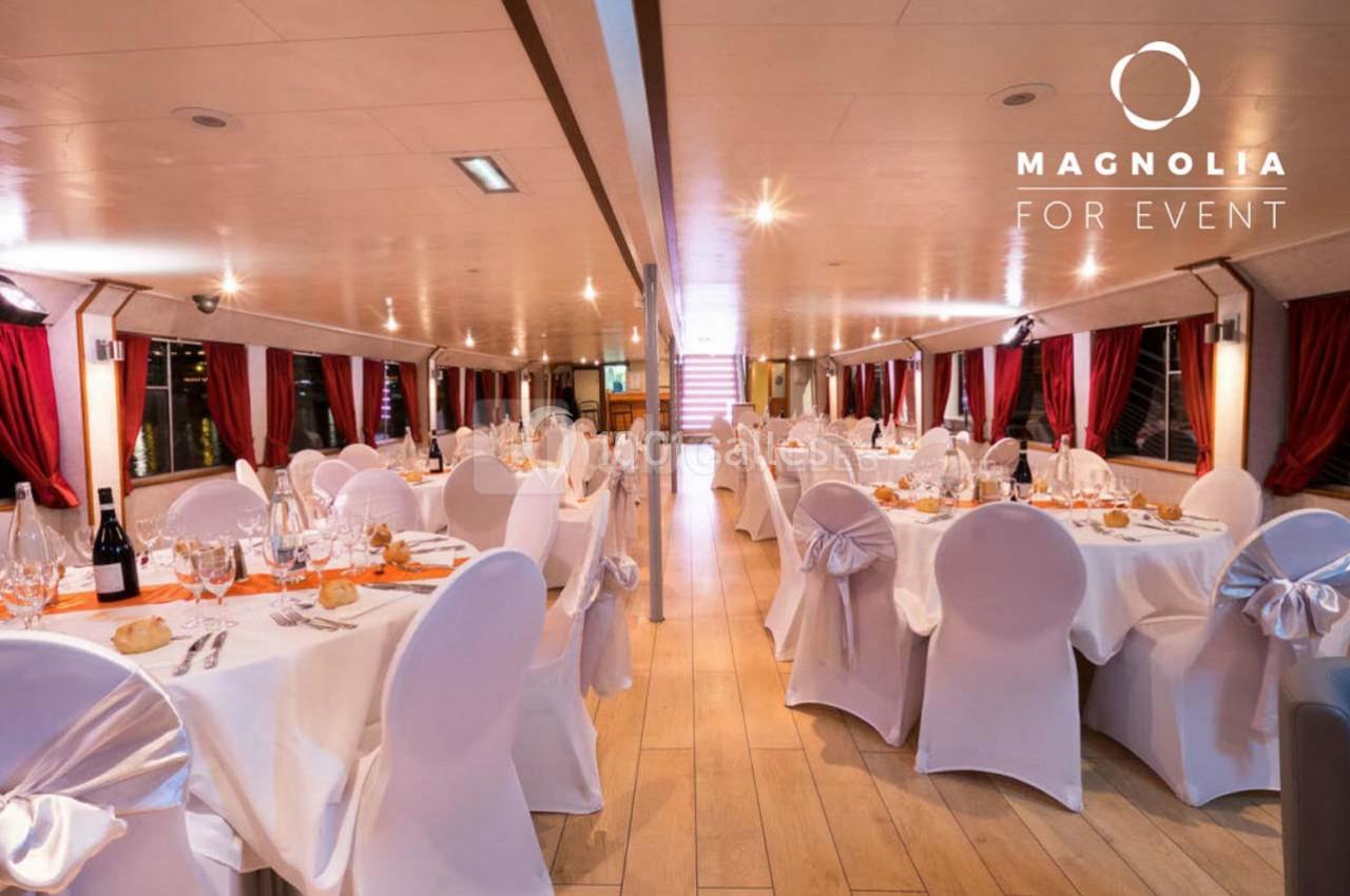 Salle de réception élégante avec tables dressées, nappes blanches et chaises décorées, éclairée par des lumières…