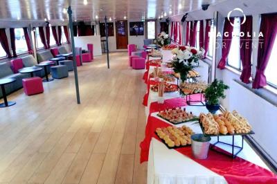 Salle de réception sur une péniche avec tables rondes dressées, rideaux rouges et buffet de desserts au premier plan.