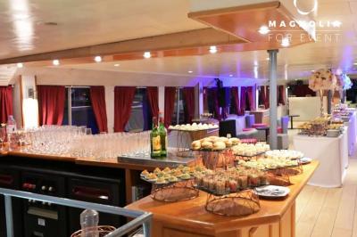 Salle de réception sur une péniche avec tables rondes dressées, rideaux rouges et buffet de desserts au premier plan.
