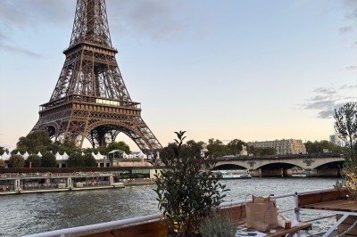 Intérieur d'un bateau avec éclairage vert, tables et chaises, vue sur une terrasse extérieure et des passants.