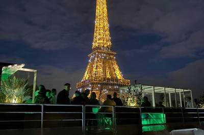 Intérieur d'un bateau avec éclairage vert, tables et chaises, vue sur une terrasse extérieure et des passants.