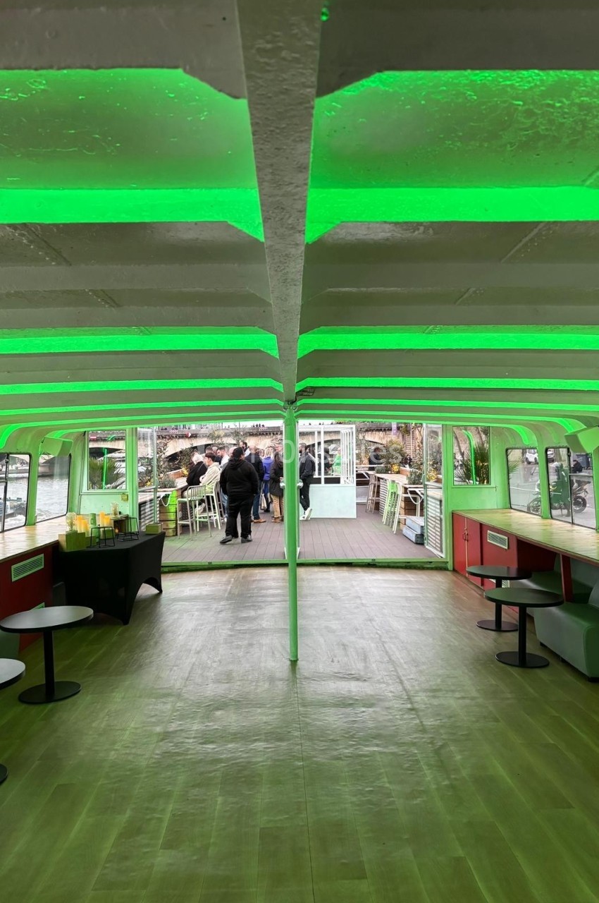 Intérieur d'un bateau avec éclairage vert, tables et bancs, vue sur une terrasse extérieure avec des personnes.