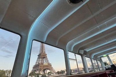 Intérieur d'un bateau avec éclairage vert, tables et chaises, vue sur une terrasse extérieure et des passants.