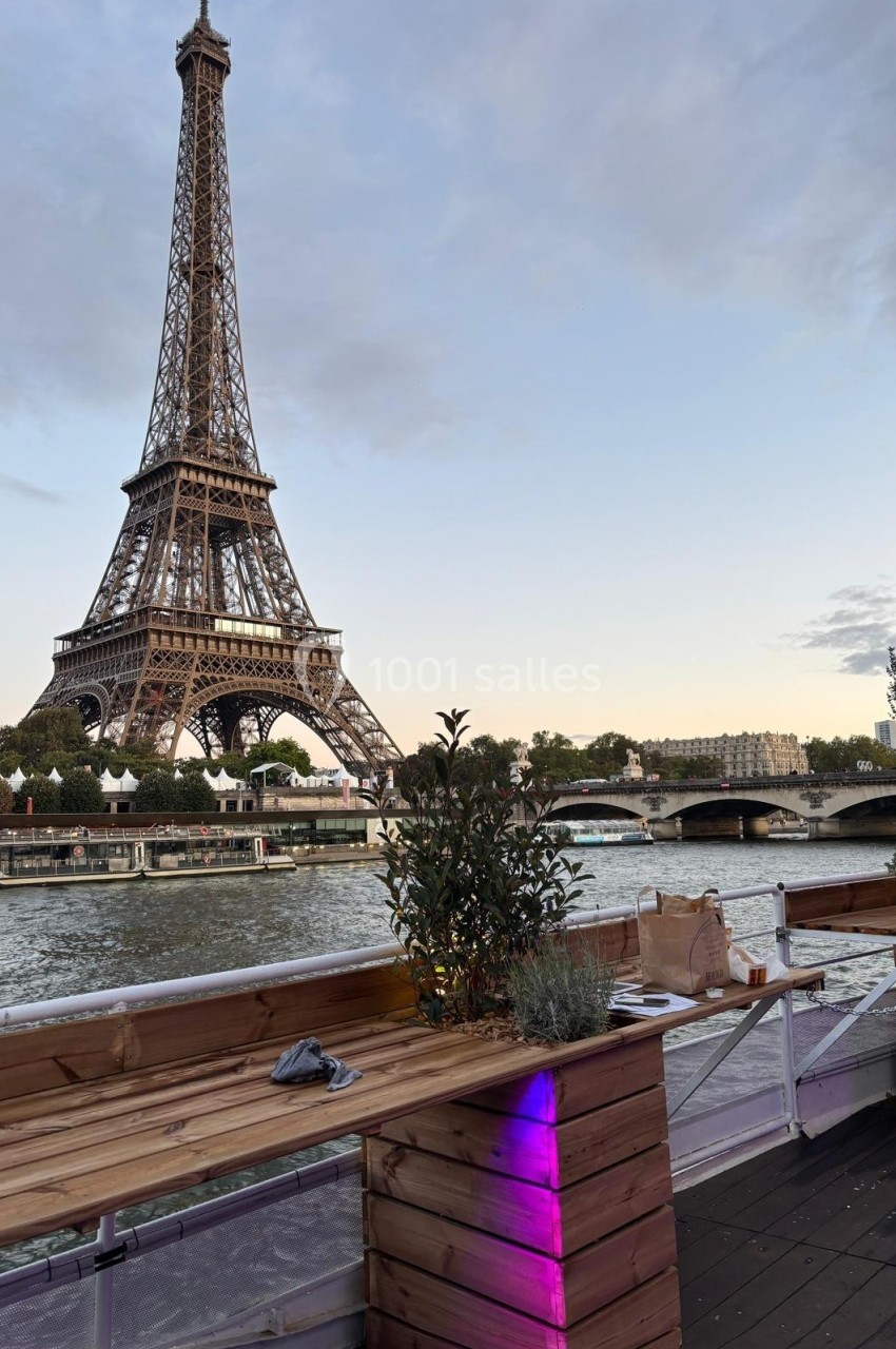 Vue de la tour Eiffel depuis une terrasse en bois au bord de la Seine, avec des plantes et des sacs posés sur une table.