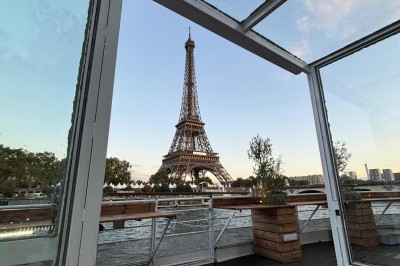 Intérieur d'un bateau avec éclairage vert, tables et chaises, vue sur une terrasse extérieure et des passants.
