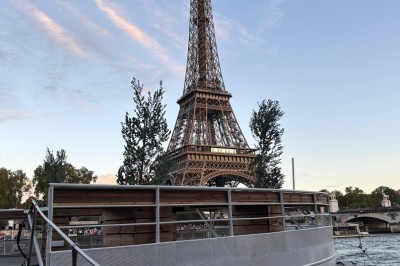 Intérieur d'un bateau avec éclairage vert, tables et chaises, vue sur une terrasse extérieure et des passants.