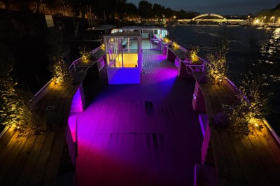 Intérieur d'un bateau avec éclairage vert, tables et chaises, vue sur une terrasse extérieure et des passants.