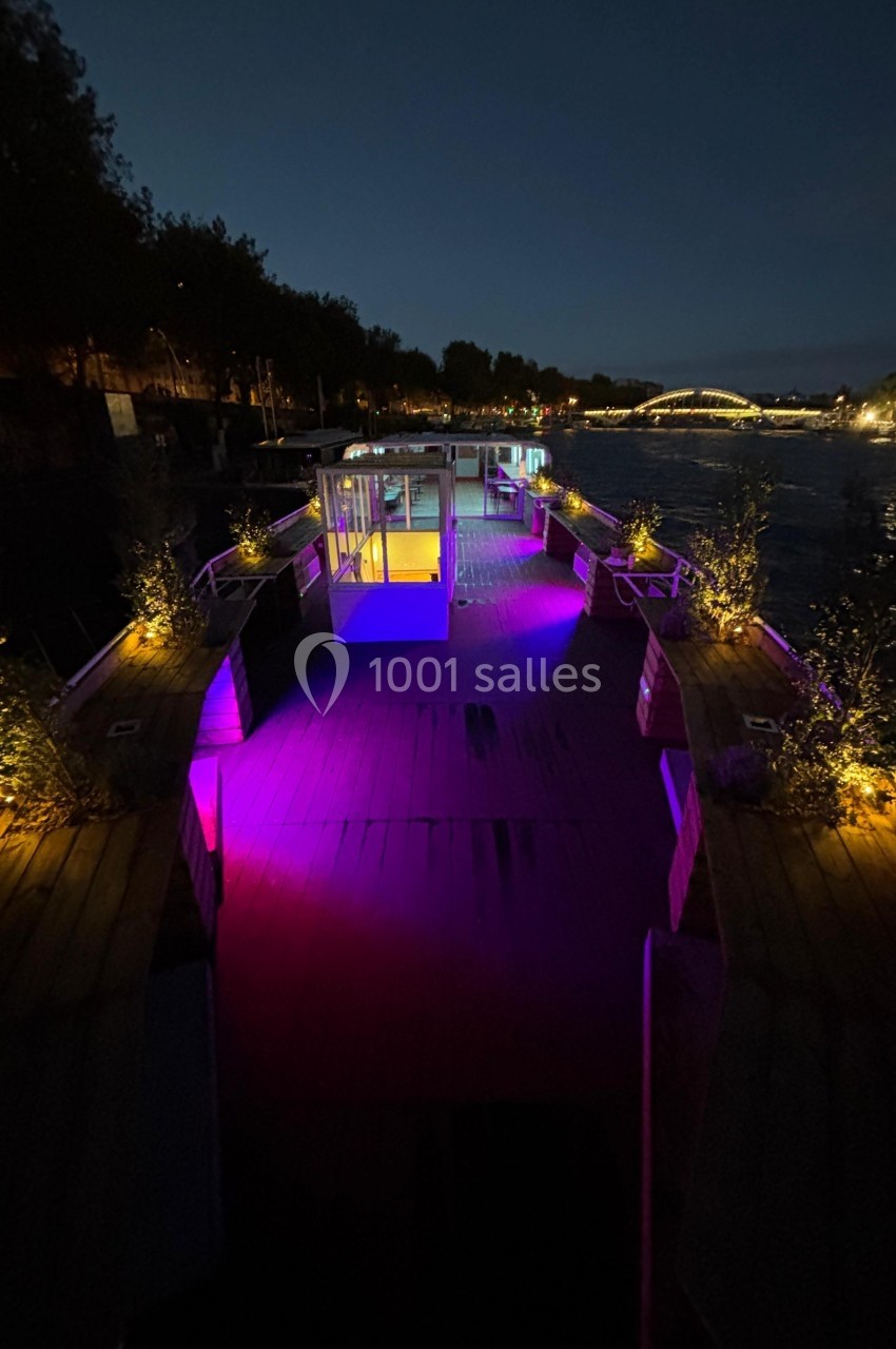 Pont d'une péniche éclairée par des lumières colorées, amarrée au bord d'une rivière en soirée.