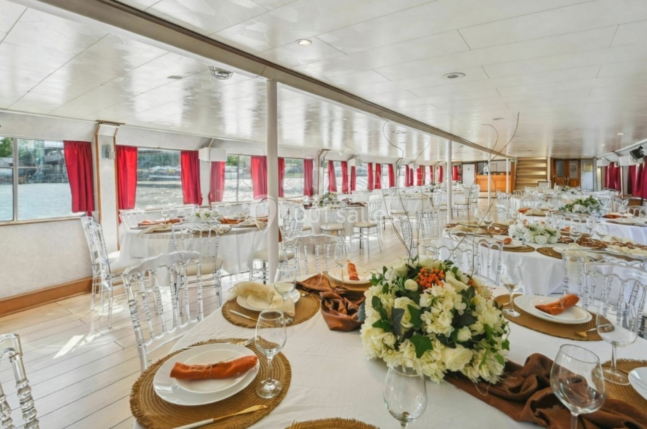 Salle de réception lumineuse sur un bateau, avec tables dressées, nappes blanches et décoration florale élégante.