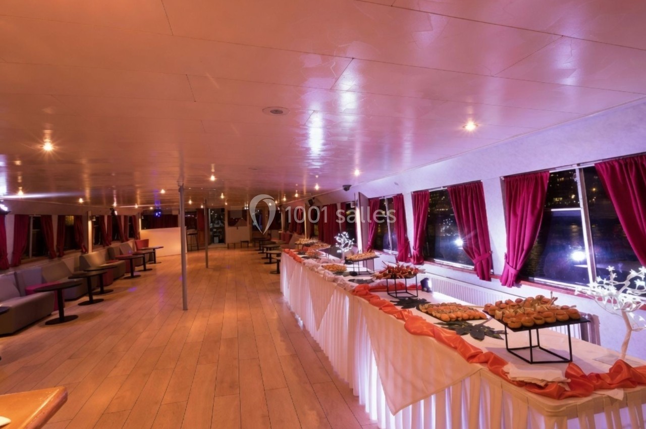 Salle de réception avec buffet dressé, éclairage chaleureux, grandes fenêtres et rideaux rouges.