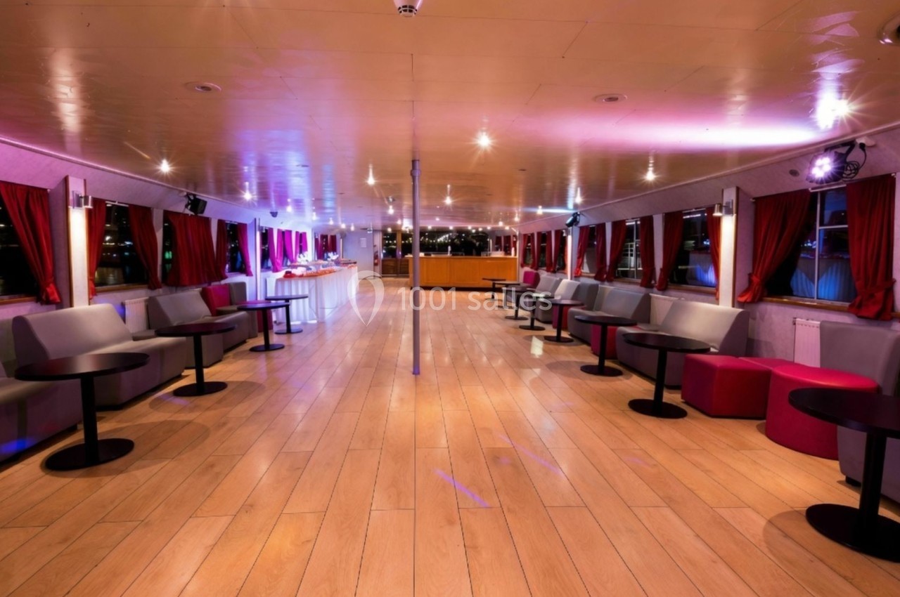 Intérieur d'un bateau avec un sol en bois, des banquettes, des tables basses et des rideaux rouges, éclairage tamisé.