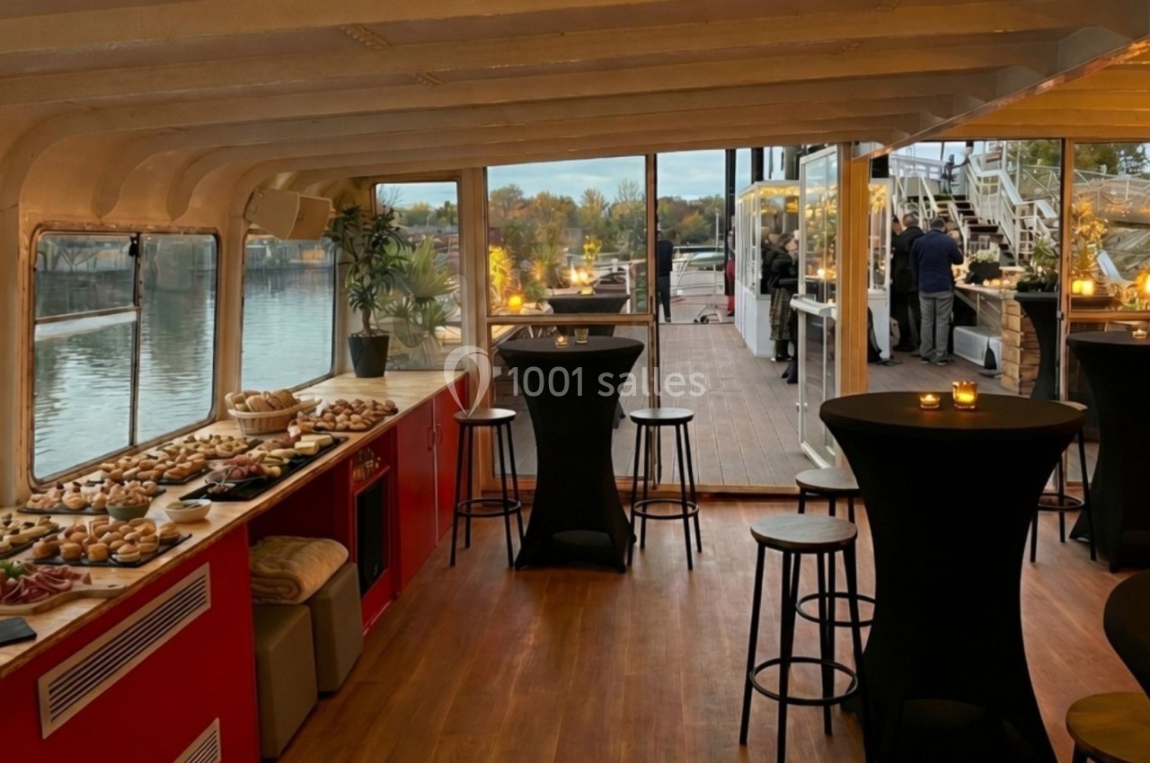 Salle intérieure d'un bateau aménagée pour un événement, avec buffet, tables hautes et vue sur une terrasse extérieure.